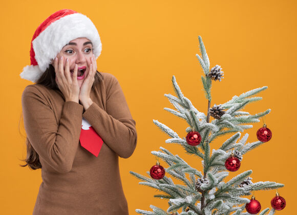 帽子惊讶的年轻漂亮女孩戴着圣诞帽打着领带站在圣诞树旁把手放在隔离在橙色墙上的脸颊上脸颊领带惊喜