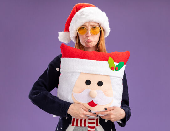 眼镜悲伤的年轻漂亮女孩穿着圣诞毛衣戴着圣诞帽戴着眼镜抱着圣诞枕头孤立在紫色的墙上毛衣戴着抱着