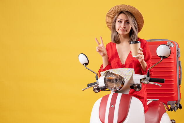 制造正面图：身穿红色连衣裙的年轻女士手持咖啡杯 在轻便摩托车旁制作胜利标志持有轻便摩托车旅游