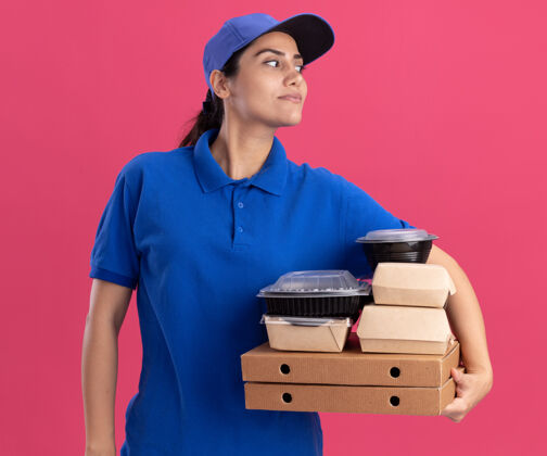 穿着高兴地看着身边穿着制服 戴着帽子的年轻送货女孩把食物容器放在隔离在粉红色墙上的比萨饼盒上年轻人人容器