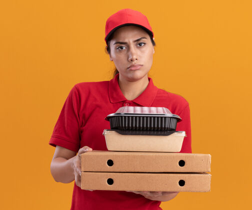 盒子一个穿着制服 戴着帽子 拿着披萨盒和食物容器的年轻送货女孩被隔离在橙色的墙上不愉快食物脸