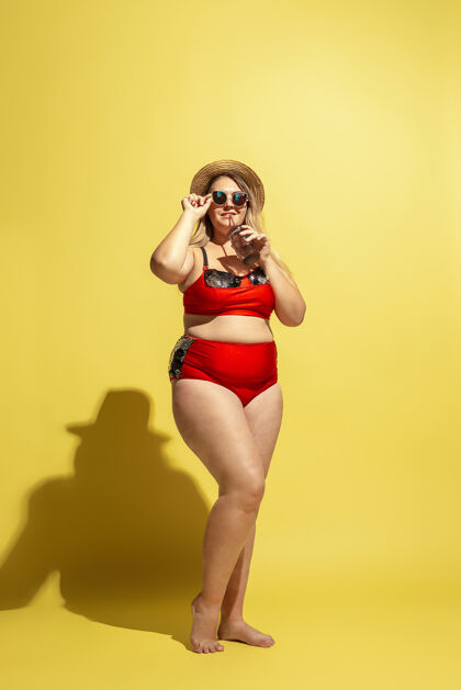 专业年轻的白人大码女模特在黄墙上准备度假穿着红色泳衣 戴着帽子和太阳镜的女人运动员努力混合