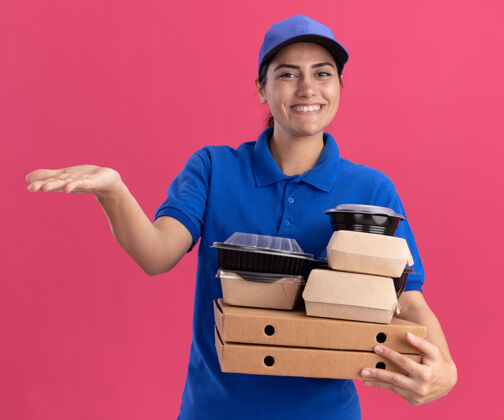 制服面带微笑的年轻送货员身穿制服 头戴鸭舌帽 手拿食物容器放在披萨盒上 手指放在隔离在粉红色墙上的一侧食物送货人