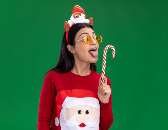 手杖年轻的白人女孩戴着圣诞老人的头带和毛衣 戴着眼镜 看着传统的圣诞糖果手杖 舌头正准备舔它 隔离在绿色墙壁上 留着复制空间穿女孩圣诞老人