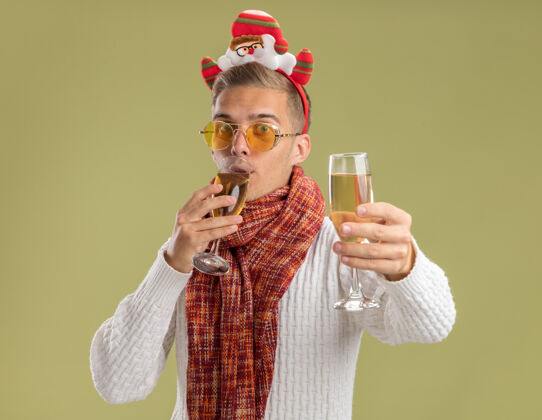 小伙子给人印象深刻的年轻帅哥戴着圣诞老人的头带和围巾 看起来拿着两杯香槟 喝着一杯 伸开另一杯对着橄榄绿墙上的相机伸展走向围巾
