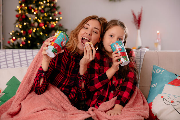 请享受高兴的母女俩捧着纸杯 吃着饼干 坐在铺着毯子的沙发上 在家里享受圣诞节的时光坐着房子女儿