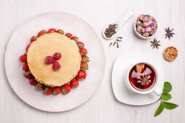 顶部俯瞰美味的草莓煎饼和一杯白底茶水果馅饼蛋糕饼干甜浆果茶杯子盘子
