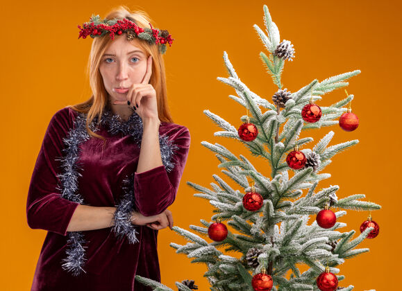 不愉快站在圣诞树旁的年轻漂亮女孩 身穿红色连衣裙 脖子上戴着花环 手放在脸颊上 被隔离在橙色的墙上花圈手女孩