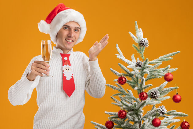 圣诞老人微笑着的年轻帅哥戴着圣诞帽 打着圣诞老人的领带 站在装饰好的圣诞树旁 手里拿着一杯香槟 看上去是一只空手孤立在橙色的墙上帽子抱着圣诞