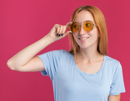 女孩高兴的年轻红发姜姑娘带着雀斑在太阳眼镜边看太阳粉红色壁板
