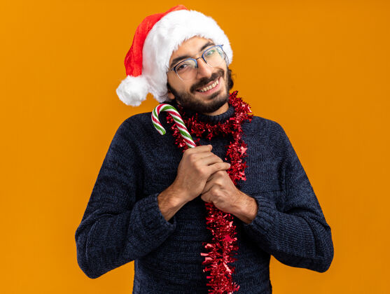 圣诞节年轻帅哥戴着圣诞帽 脖子上戴着花环 手里拿着圣诞糖果 孤零零地站在橙色的墙上年轻人糖果花环