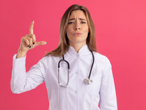 人年轻的女医生穿着医用长袍 听诊器显示出粉红色墙上孤立的大小脸表情长袍
