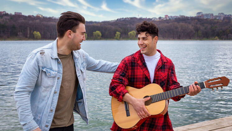 树两个年轻的朋友在公园的湖边唱歌和弹吉他年轻生活方式胡同