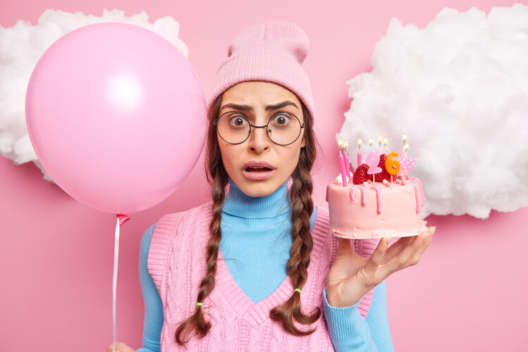庆典女人庆祝她的26岁生日拿着蛋糕点燃蜡烛和充气气球戴着粉色帽子高领毛衣大圆眼镜站在室内凝视场合女人
