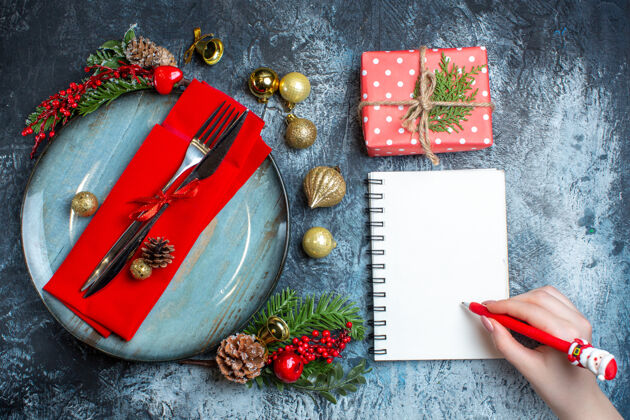 笔记本上图：手拿一支笔放在螺旋笔记本和餐具旁的礼品盒上背景餐具套装礼品