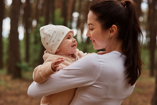 人类美丽的黑发女子穿着白色衣服在户外摆姿势 手里抱着婴儿 用极大的爱看着女儿 在森林里玩耍生活女孩家庭