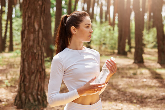 健康健身美女 黑发马尾 手拿一瓶水 望着别处 在森林里锻炼后摆出姿势和谐运动自然