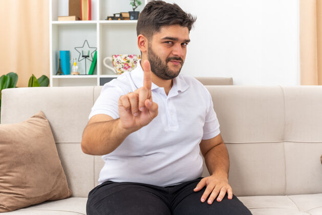 休闲一个穿着休闲服的年轻人 自信的表情 展示着食指警告的手势 坐在明亮的客厅里的沙发上手指表情表演