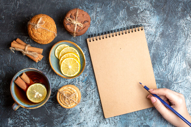 深色上图是新鲜的柠檬和一杯红茶 各种各样的肉桂饼干叠在一起 手里拿着一支笔 在深色的背景上写着一本笔记本扣件纸笔记本