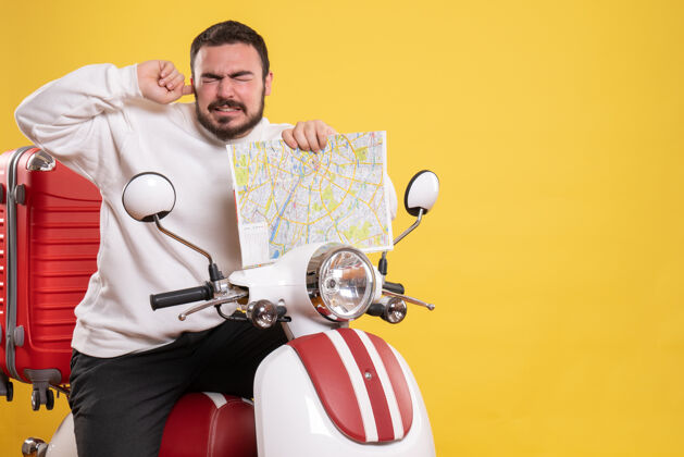 摩托车前视图的麻烦男子坐在摩托车上 手提箱上拿着地图痛苦的耳朵在孤立的黄色背景手提箱地图抱着