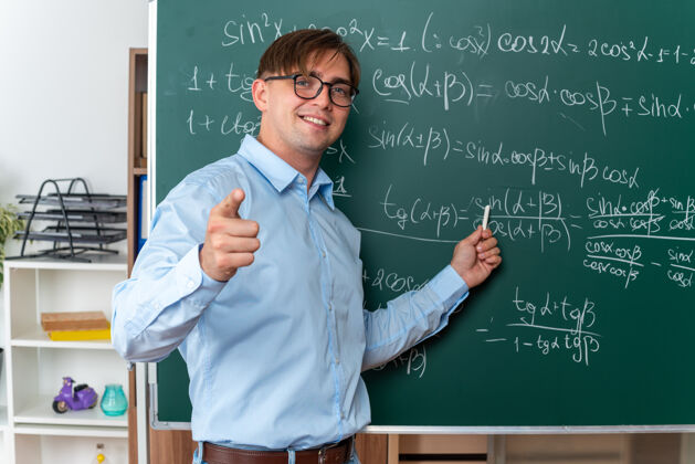 年轻年轻的男老师戴着眼镜 用粉笔讲课 快乐而积极地微笑着站在教室里数学公式的黑板旁积极解释课