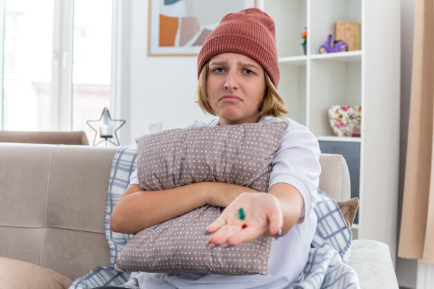 女人不健康的年轻女子 戴着暖和的帽子 拿着毯子 感觉不舒服 生病 患感冒和流感 抱着枕头和药片 坐在客厅里的沙发上忧心忡忡不适温暖流感