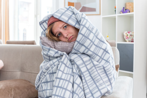 生病不健康的年轻女子 戴着暖和的帽子 裹着毯子 坐在明亮的客厅里的沙发上 感觉不舒服 生病 看起来很担心 患流感和感冒不适光沙发
