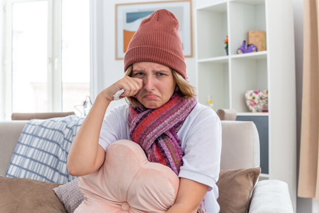 摩擦不健康的年轻女士戴着暖和的帽子 戴着围巾 看起来不舒服 病了 拿着纸巾 患着感冒和流感 哭着 使劲揉眼睛 坐在明亮的客厅沙发上生病流感年轻