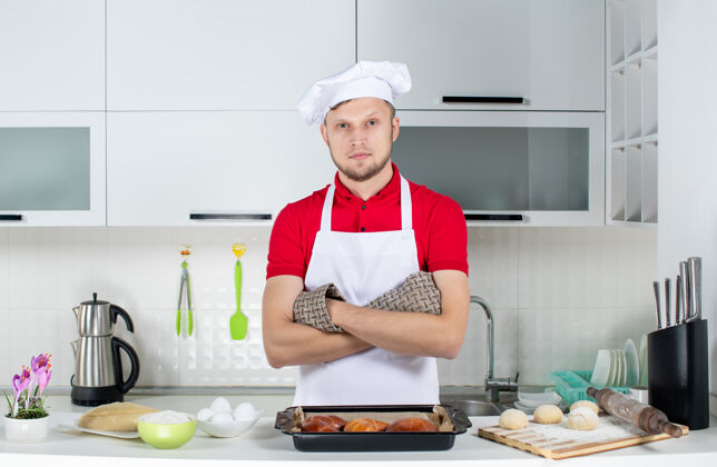 持有人俯视图：白色厨房里 年轻的厨师戴着支架站在桌子后面 上面放着糕点和鸡蛋磨碎器微笑糕点立场