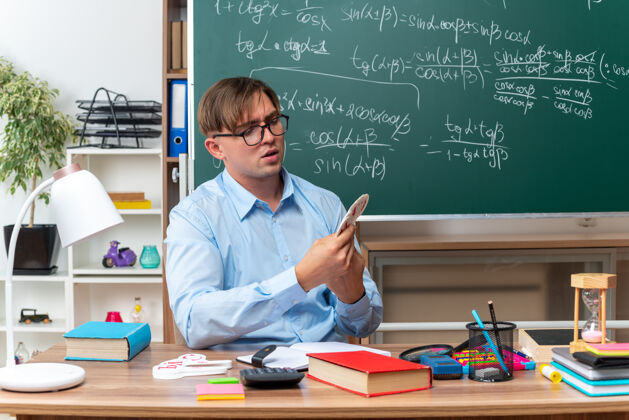 黑板年轻的男老师戴着眼镜上课 看起来很自信 坐在课桌旁 教室的黑板前放着书和笔记课桌教室穿