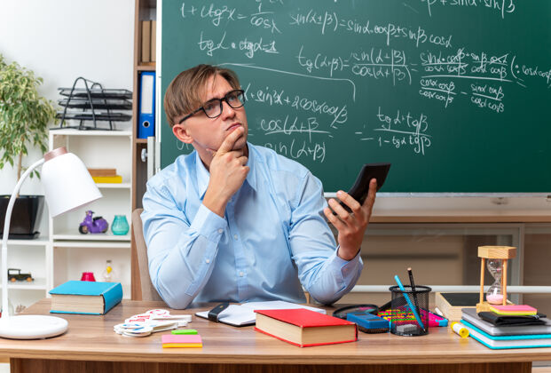 学校年轻的男老师戴着眼镜用智能手机打字 神情困惑地坐在课桌旁 教室的黑板前放着书和笔记穿打字教室