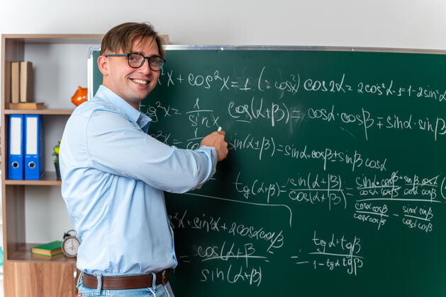 课戴着眼镜的年轻男老师在教室里兴高采烈地微笑着站在黑板旁讲解数学公式穿教室积极