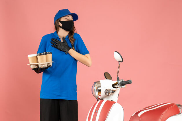 人年轻的梦幻信使女孩戴着医用口罩手套站在摩托车旁 手里拿着粉色背景上的咖啡小蛋糕医疗手套年轻