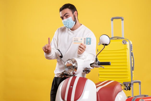 黄色旅行概念的俯视图 戴着医用面罩的微笑的家伙站在摩托车旁边 黄色的手提箱放在上面 拿着车票朝上抱着快乐人