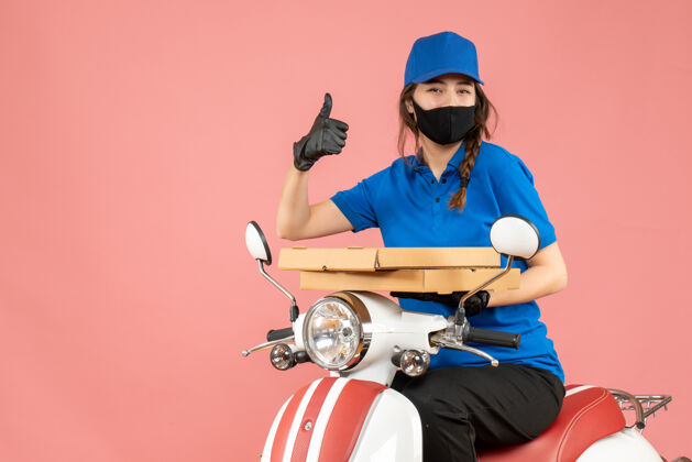 手套俯视图：微笑的女快递员戴着医用口罩和手套坐在滑板车上传递订单 在柔和的桃色上做着ok的手势桃摩托车头盔