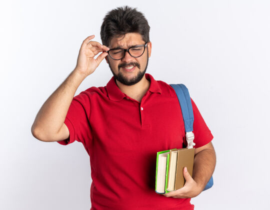 姿势年轻的留着胡子的学生 穿着红色马球衫 背着背包 拿着笔记本 戴着眼镜 快乐地站在白墙上背包穿着年轻人
