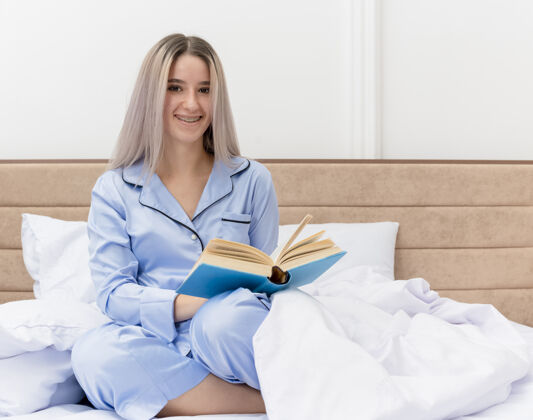 漂亮穿着蓝色睡衣的年轻漂亮女人坐在床上 带着一本书微笑着 一张幸福的脸躺在卧室里卧室女人床