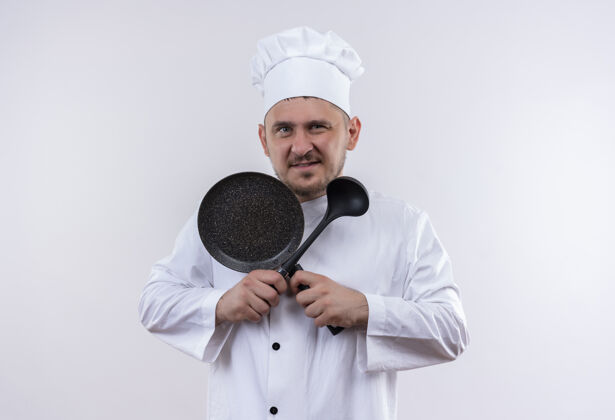 英俊自信的年轻帅哥厨师身着厨师制服 手拿煎锅和勺子 隔离在白墙上薯条勺子锅