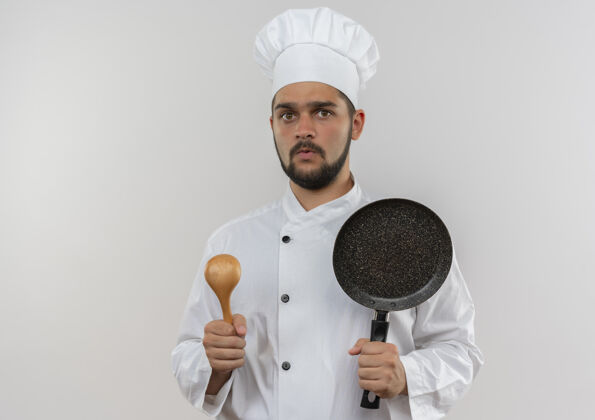 平底锅印象深刻的年轻男厨师在厨师制服举行汤匙和煎锅隔离在白色的墙壁与复制空间厨师制服印象
