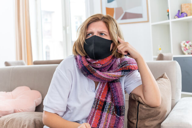 周围不健康的年轻女子 脖子上围着暖和的围巾 戴着面膜 感觉不舒服 生病 患流感和感冒 坐在客厅的沙发上 神志不清困惑沙发面部