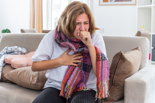 围巾不健康的年轻女子围着暖和的围巾坐在客厅的沙发上咳嗽 感觉很可怕病毒沙发咳嗽