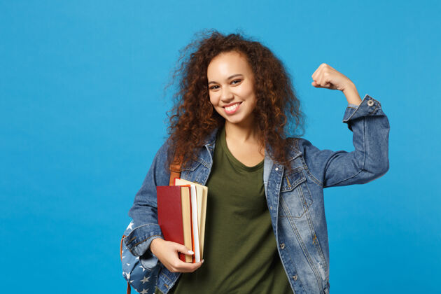 运动穿着牛仔服 背着书包的年轻女学生把书孤立地放在蓝色的墙上学习非洲手