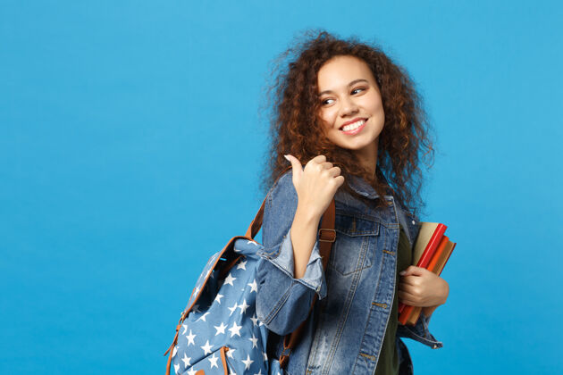 快乐穿着牛仔服 背着书包的年轻学生把书孤立地放在蓝色的墙上手微笑地方