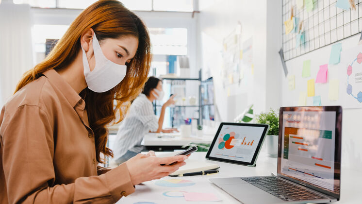 同龄人亚洲女企业家戴着口罩在新的正常情况下保持社交距离以防病毒 同时在办公室使用笔记本电脑和电话男士手机疾病