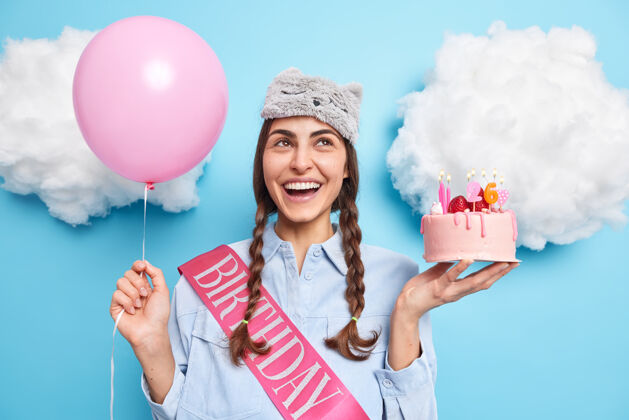 梦幻两个小辫子集中在上面的女孩表情欢快地拿着喜庆蛋糕和氦气球庆祝26岁生日在派对上等待朋友接受祝贺年轻人蛋糕睡眠面膜
