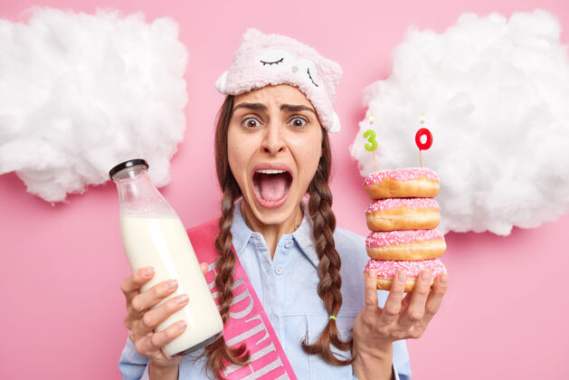 穿女性张大嘴巴独自庆祝生日大声喊叫着摆出美味的甜甜圈和牛奶隔离在粉红色的姿势美味疯狂举行