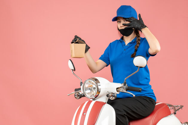 坐正面图自信的送货员戴着医用口罩和手套坐在滑板车上 在柔和的桃色背景下送货穿命令摩托车