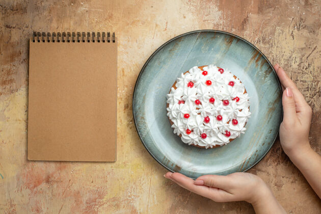 盖子上图：一只手拿着美味的蛋糕 蓝色盘子上装饰着奶油和葡萄干 彩色桌子上放着螺旋笔记本奶油盘子桌子
