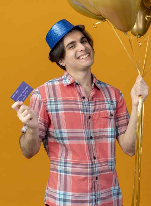 氦面带微笑的白人帅哥戴着蓝色派对帽手持氦气球和信用卡看着相机帽子男人复制