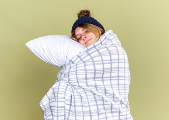 女人不健康的年轻女子戴着暖和的帽子 裹着毯子 抱着枕头 闭着眼睛微笑 站在绿色的墙壁上想睡觉站着睡眠帽子